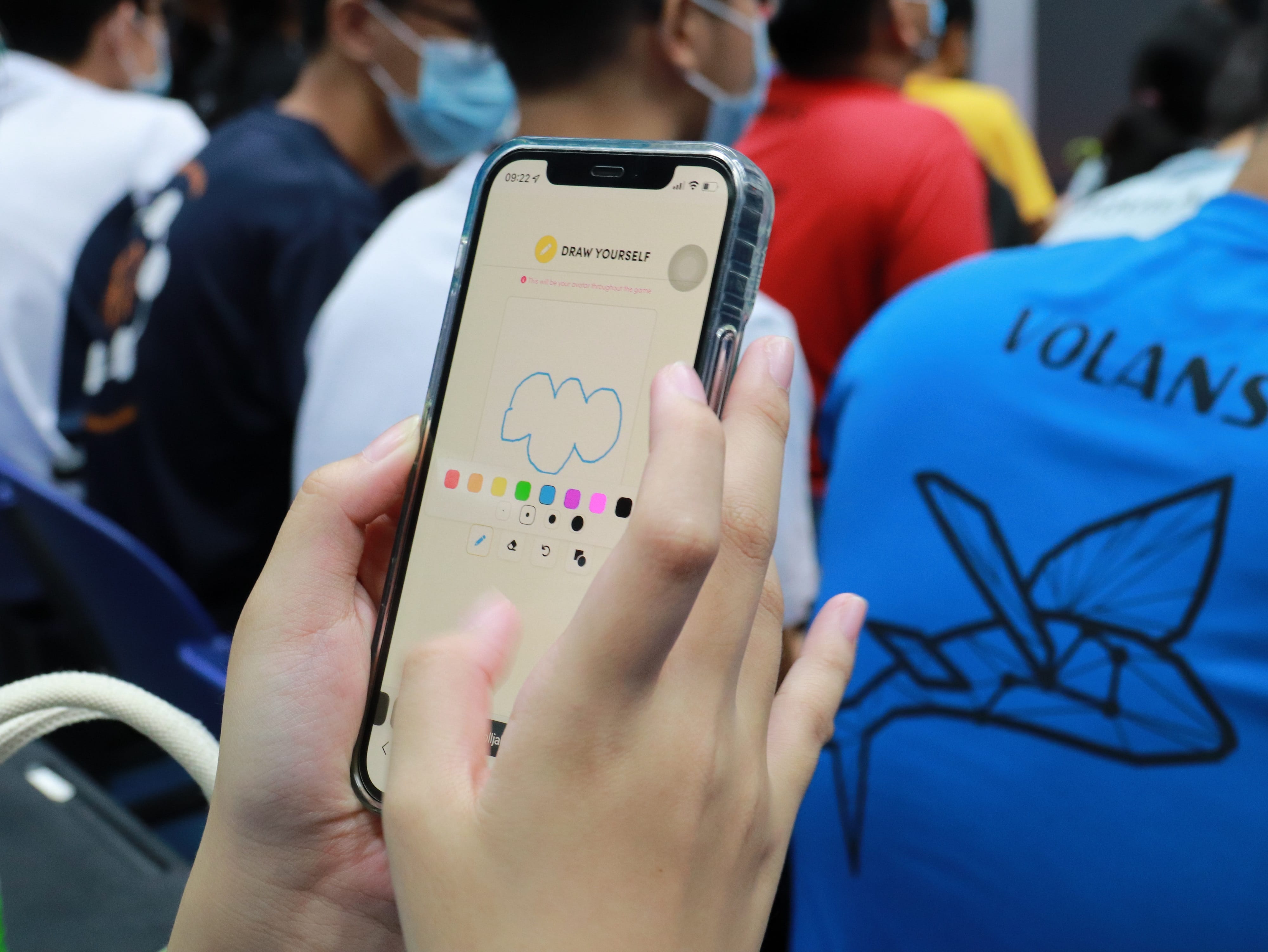 Hands holding a phone showing a drawing app with a blue sketch, surrounded by masked people in colorful shirts.
